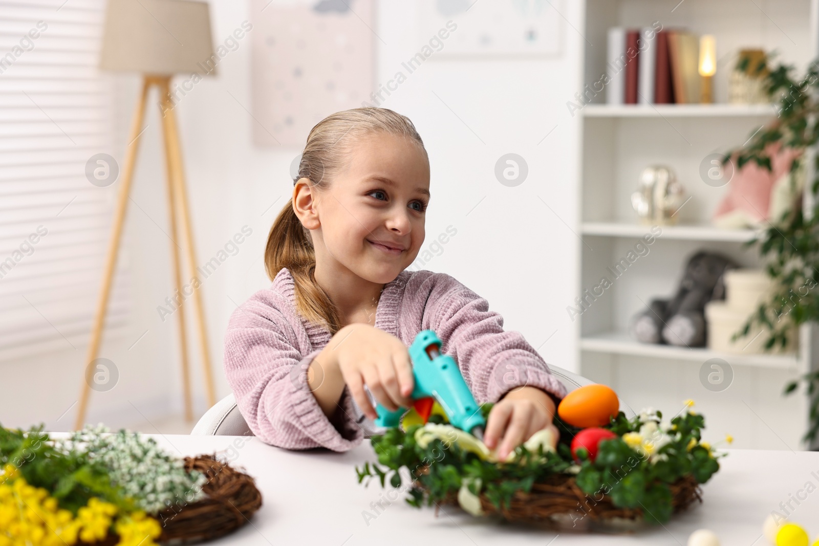 Photo of Little girl with hot glue gun creating Easter composition at table indoors