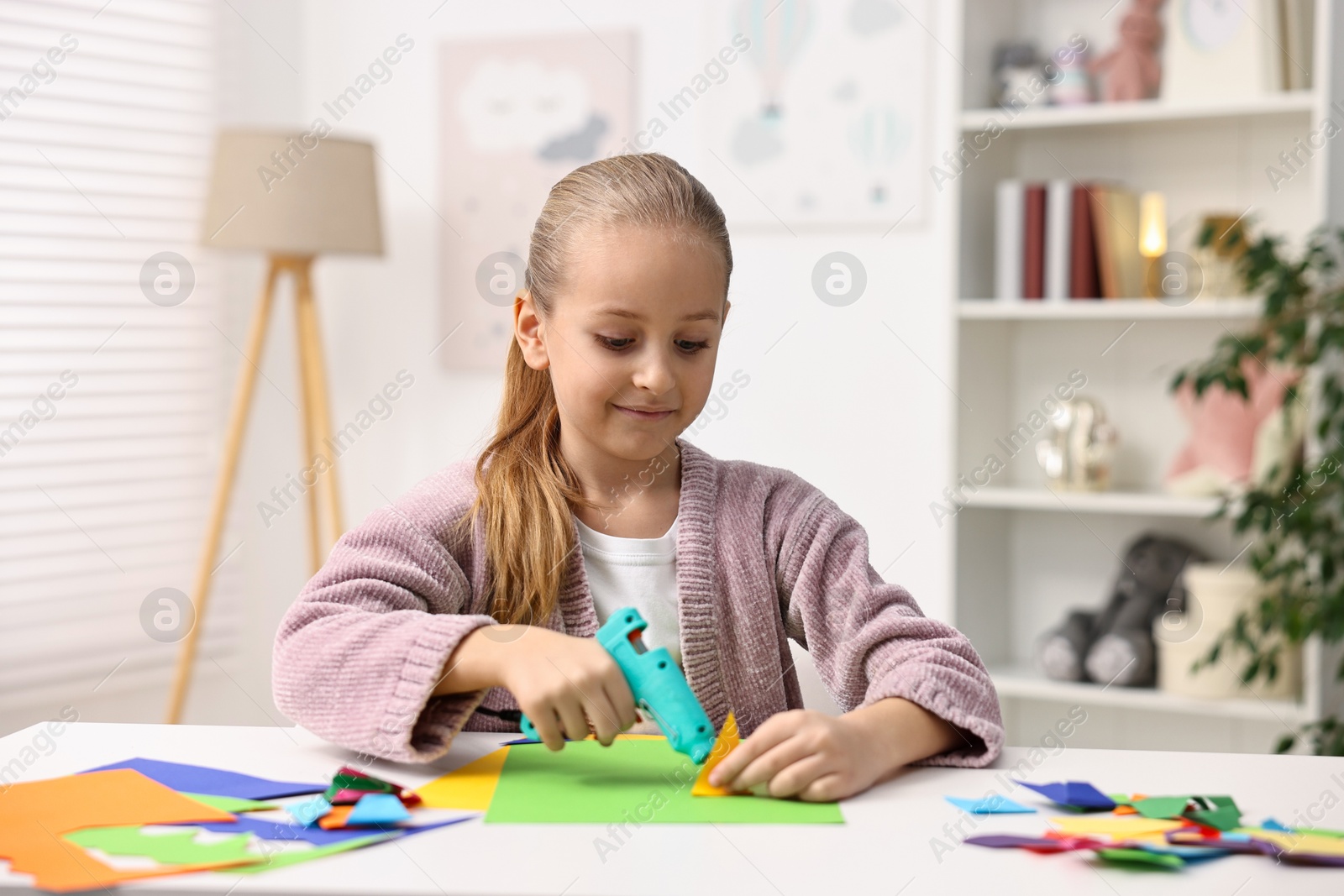 Photo of Little girl with hot glue gun and color paper making craft at table indoors