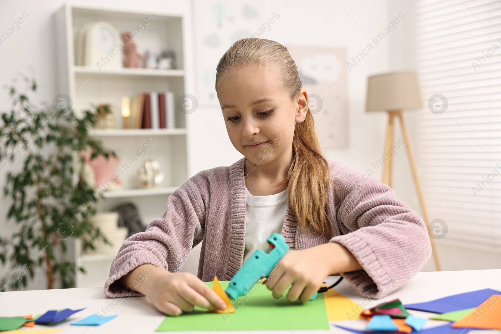 Photo of Little girl with hot glue gun and color paper making craft at table indoors