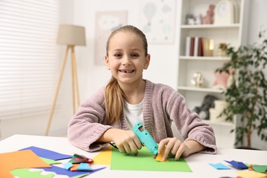 Photo of Smiling girl with hot glue gun and color paper making craft at table indoors