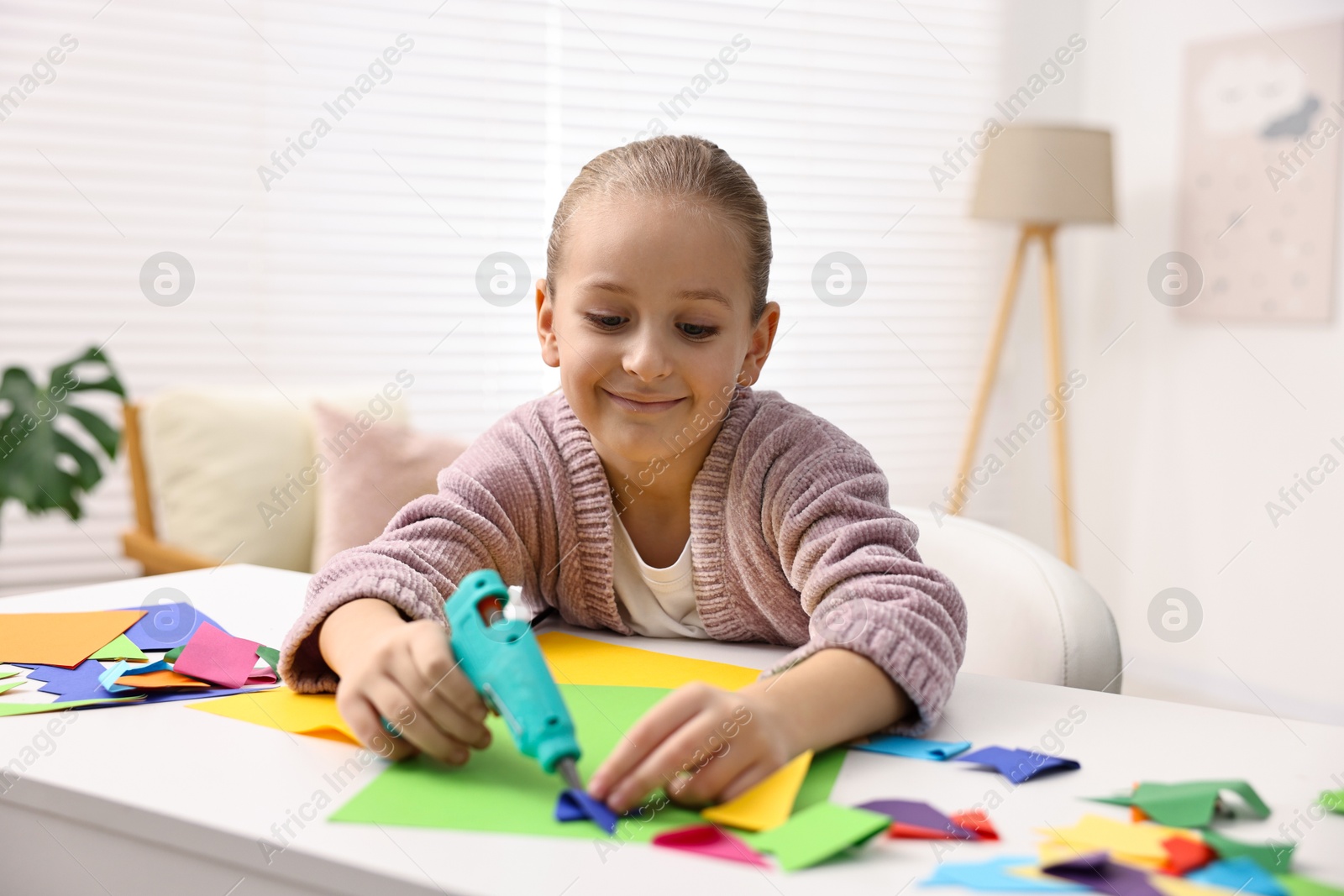 Photo of Little girl with hot glue gun and color paper making craft at table indoors