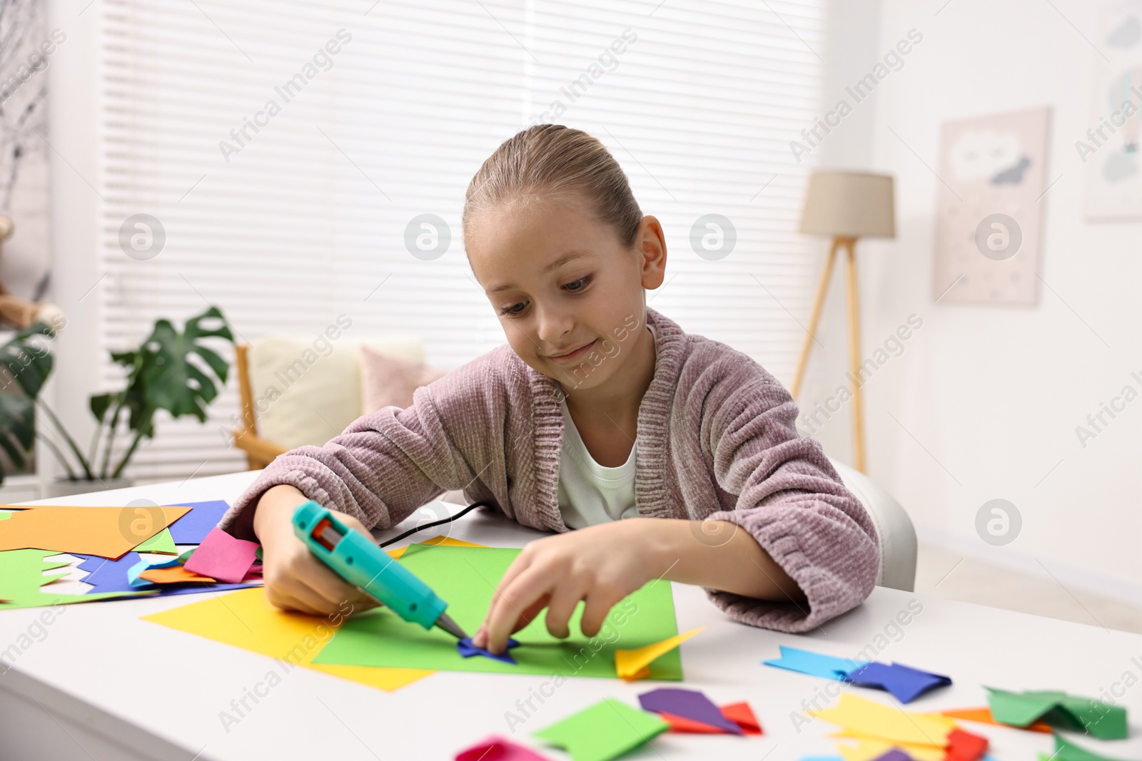 Photo of Little girl with hot glue gun and color paper making craft at table indoors