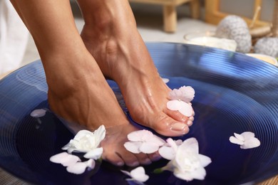 Photo of Woman soaking her feet in bowl with water and flowers on floor, closeup. Spa treatment