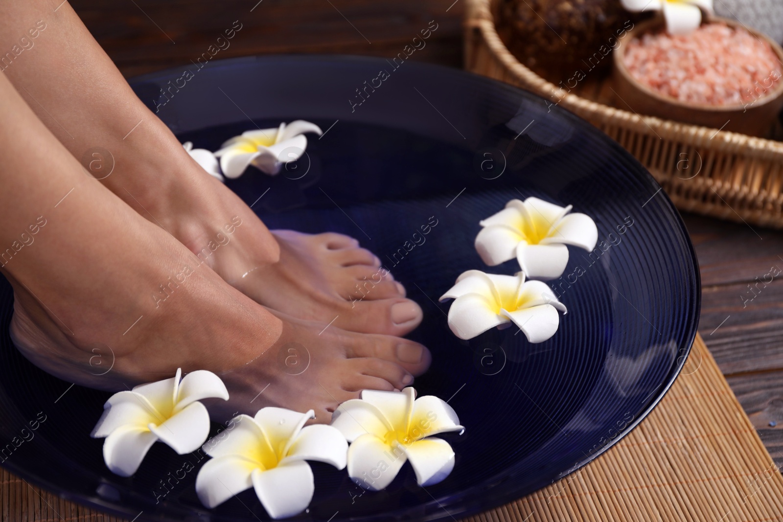 Photo of Woman soaking her feet in bowl with water and plumeria flowers on floor, closeup. Spa treatment