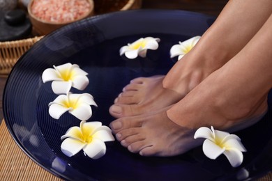 Photo of Woman soaking her feet in bowl with water and plumeria flowers on floor, closeup. Spa treatment