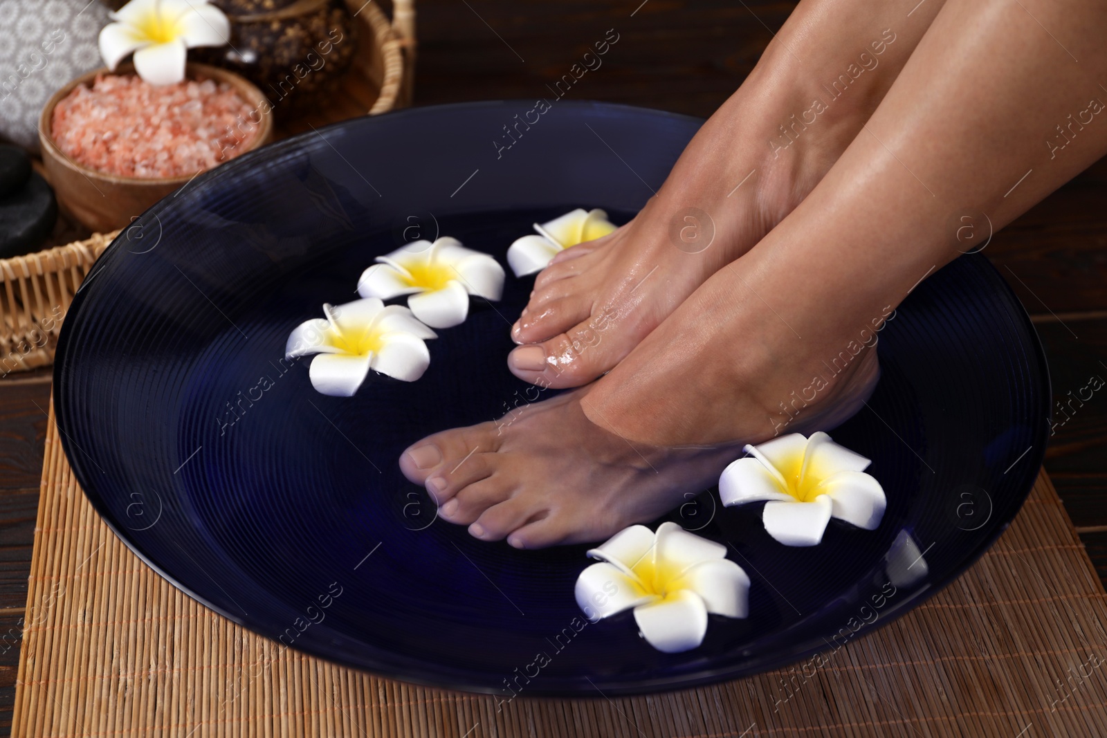 Photo of Woman soaking her feet in bowl with water and plumeria flowers on floor, closeup. Spa treatment