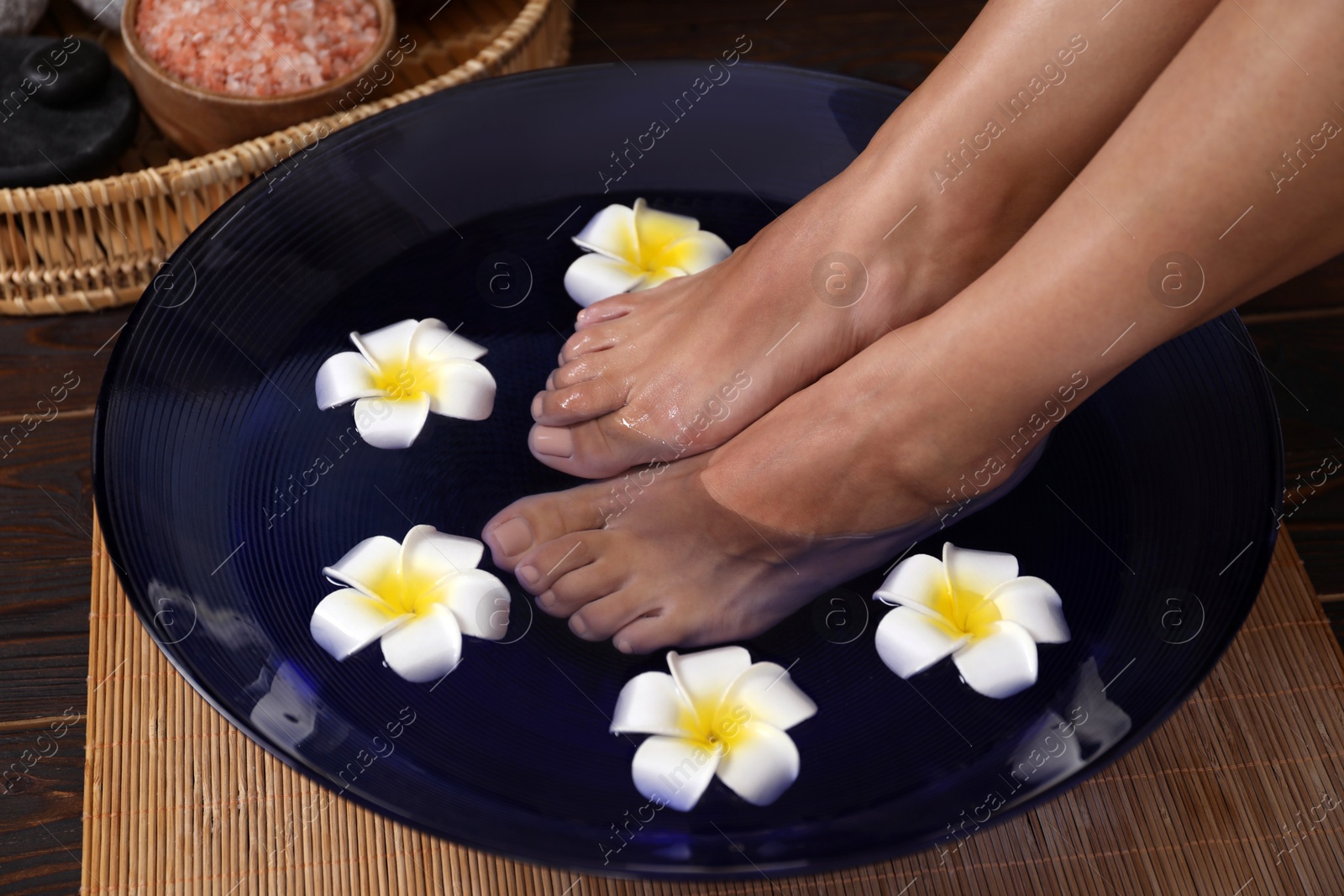 Photo of Woman soaking her feet in bowl with water and plumeria flowers on floor, closeup. Spa treatment