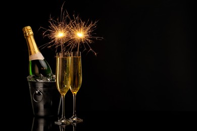 Photo of Glasses of sparkling wine with sparklers and bottle in cooler bucket on black background. Space for text