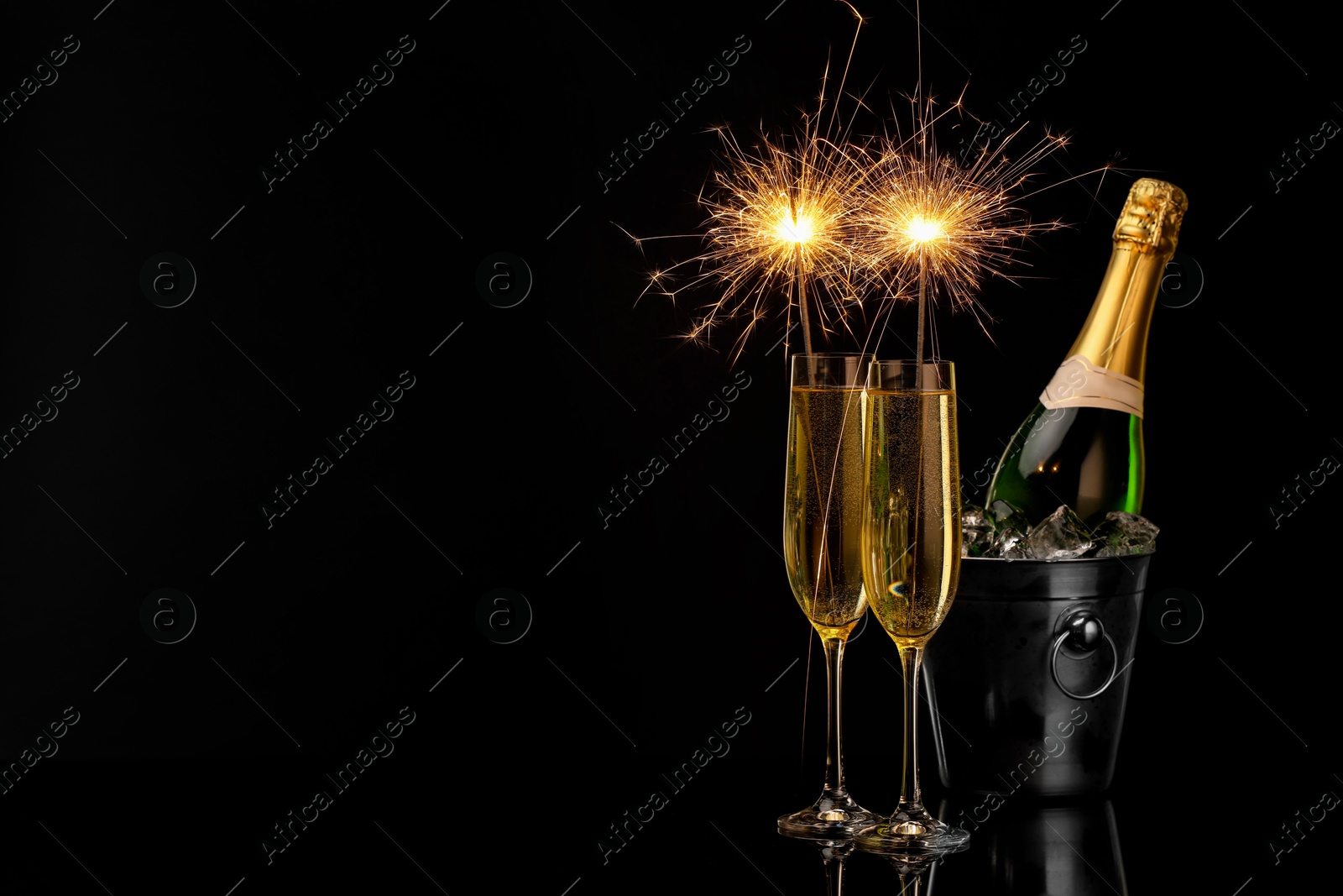 Photo of Glasses of sparkling wine with sparklers and bottle in cooler bucket on black background. Space for text