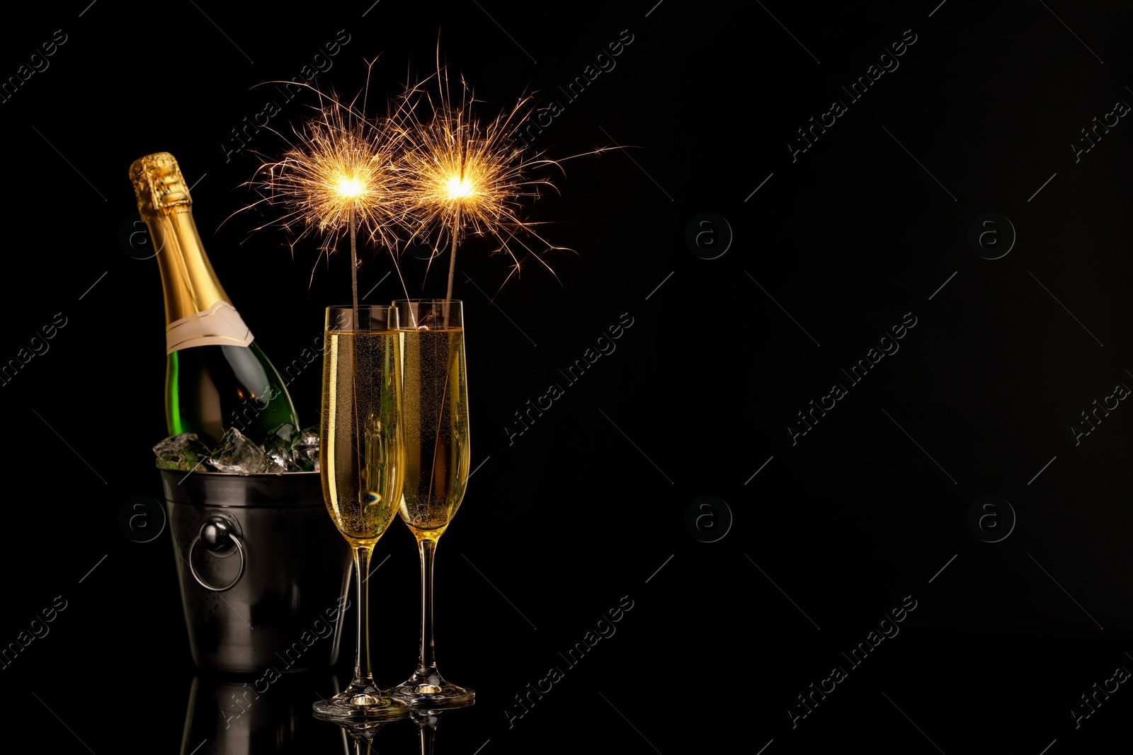 Photo of Glasses of sparkling wine with sparklers and bottle in cooler bucket on black background. Space for text