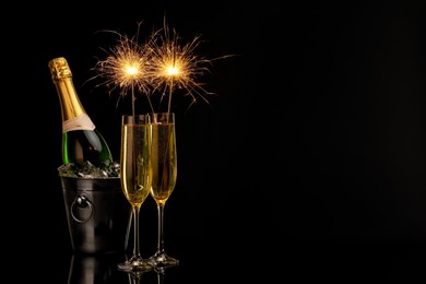 Photo of Glasses of sparkling wine with sparklers and bottle in cooler bucket on black background. Space for text