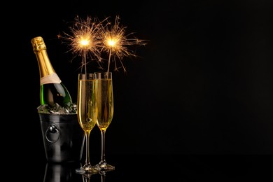 Photo of Glasses of sparkling wine with sparklers and bottle in cooler bucket on black background. Space for text