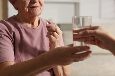 Photo of Caregiver giving glass of water to senior woman with medical pill indoors, closeup