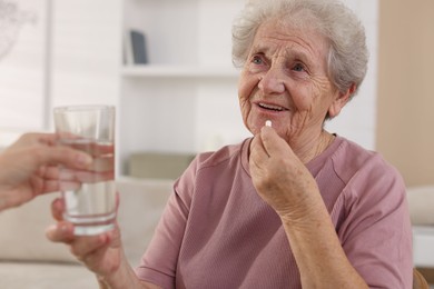 Caregiver giving glass of water to smiling senior woman with medical pill indoors, closeup