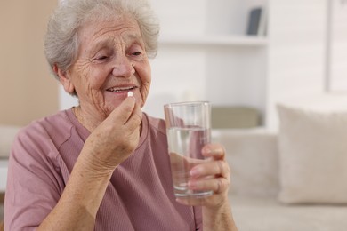 Photo of Smiling senior woman with glass of water taking pill at home. Space for text