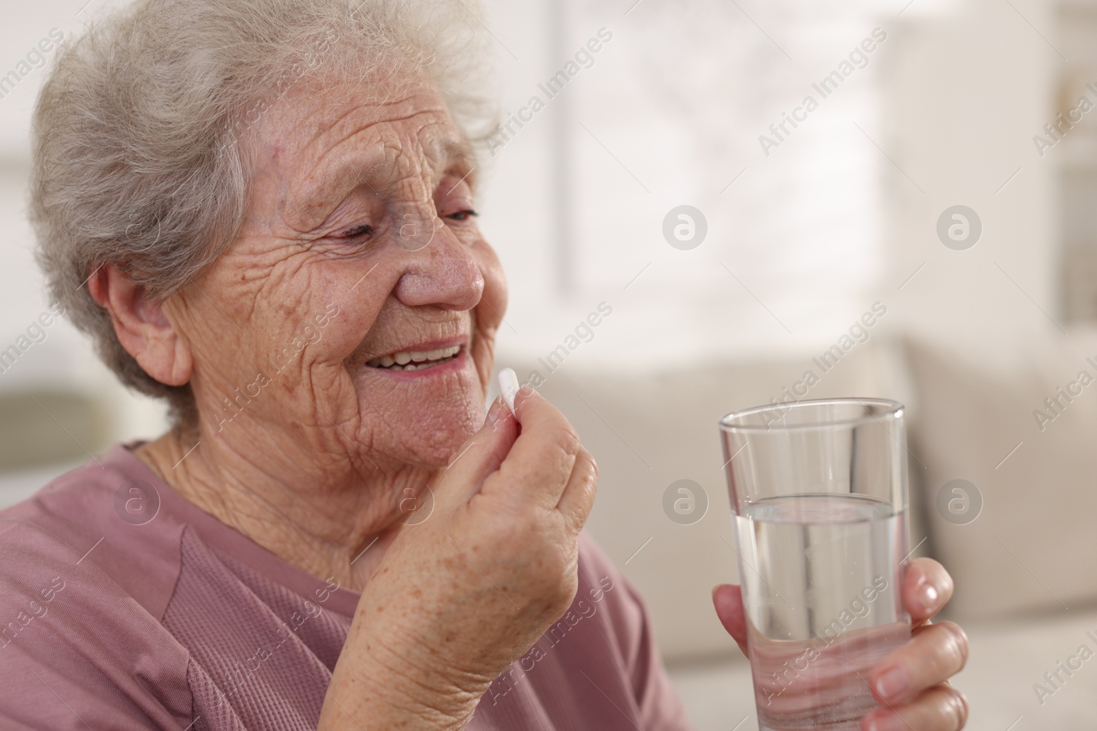 Photo of Smiling senior woman with glass of water taking pill at home, closeup