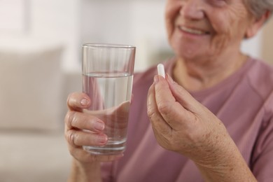 Photo of Senior woman with glass of water and pill at home, selective focus