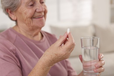 Photo of Senior woman with glass of water and pill at home, selective focus