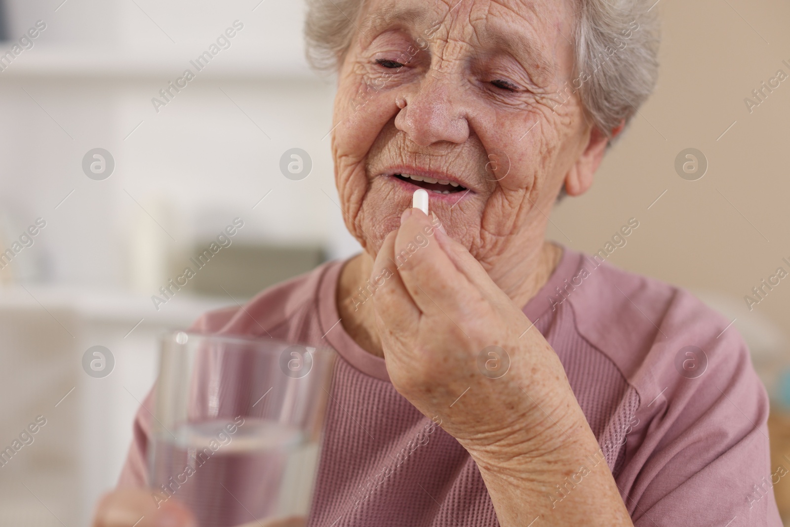 Photo of Senior woman with glass of water taking pill at home, closeup