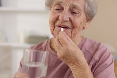 Photo of Senior woman with glass of water taking pill at home, closeup