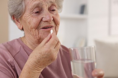 Photo of Senior woman with glass of water taking pill at home, closeup