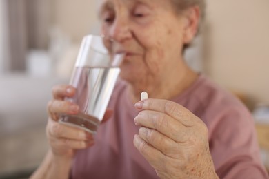 Photo of Senior woman with glass of water taking pill at home, selective focus