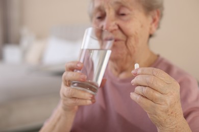 Photo of Senior woman with glass of water taking pill at home, selective focus