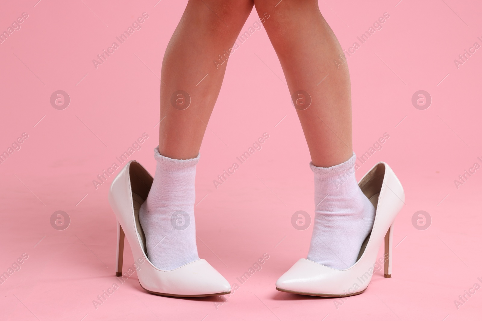 Photo of Little girl wearing oversized high heeled shoes on pink background, closeup