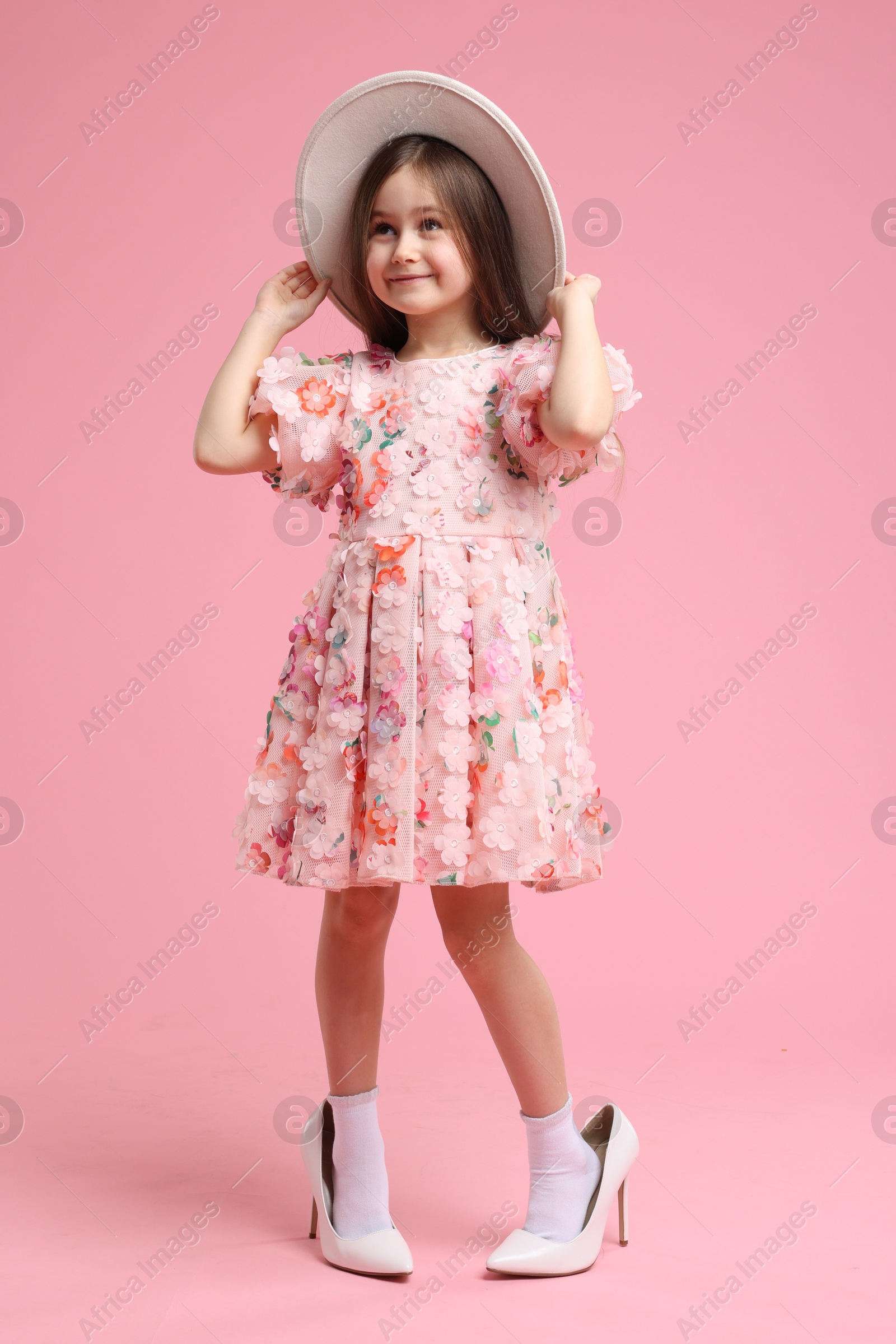 Photo of Stylish little girl wearing oversized high heeled shoes on pink background