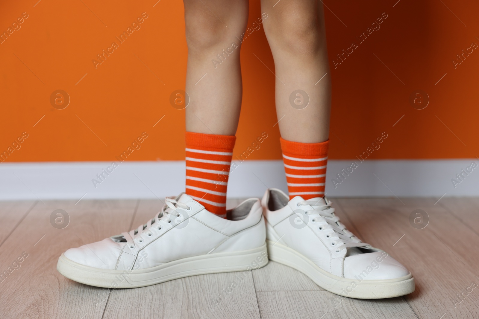 Photo of Child wearing oversized sneakers near orange wall indoors, closeup