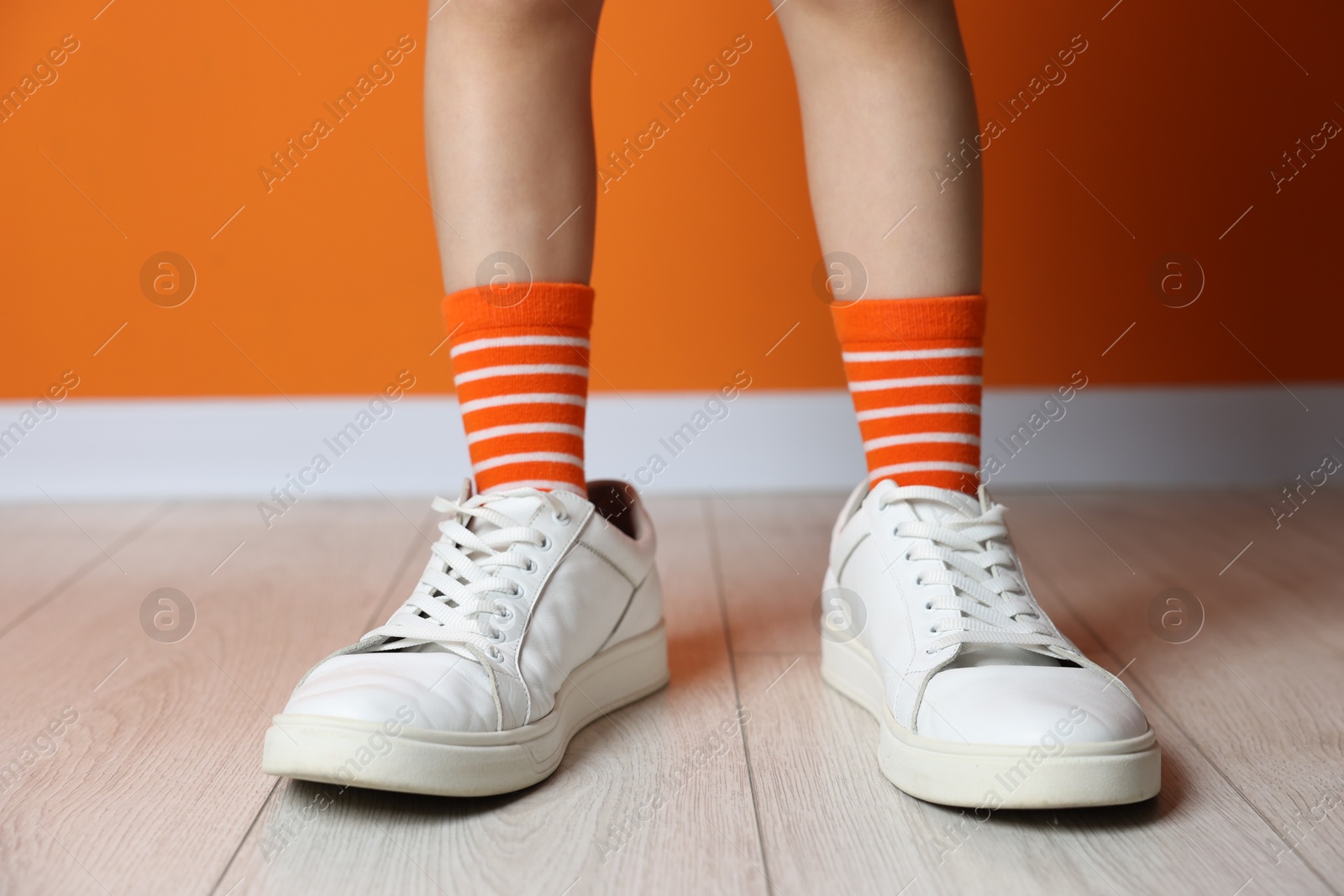 Photo of Child wearing oversized sneakers near orange wall indoors, closeup