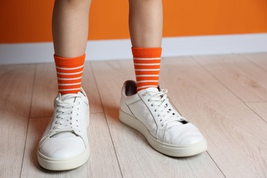 Photo of Child wearing oversized sneakers near orange wall indoors, closeup. Space for text