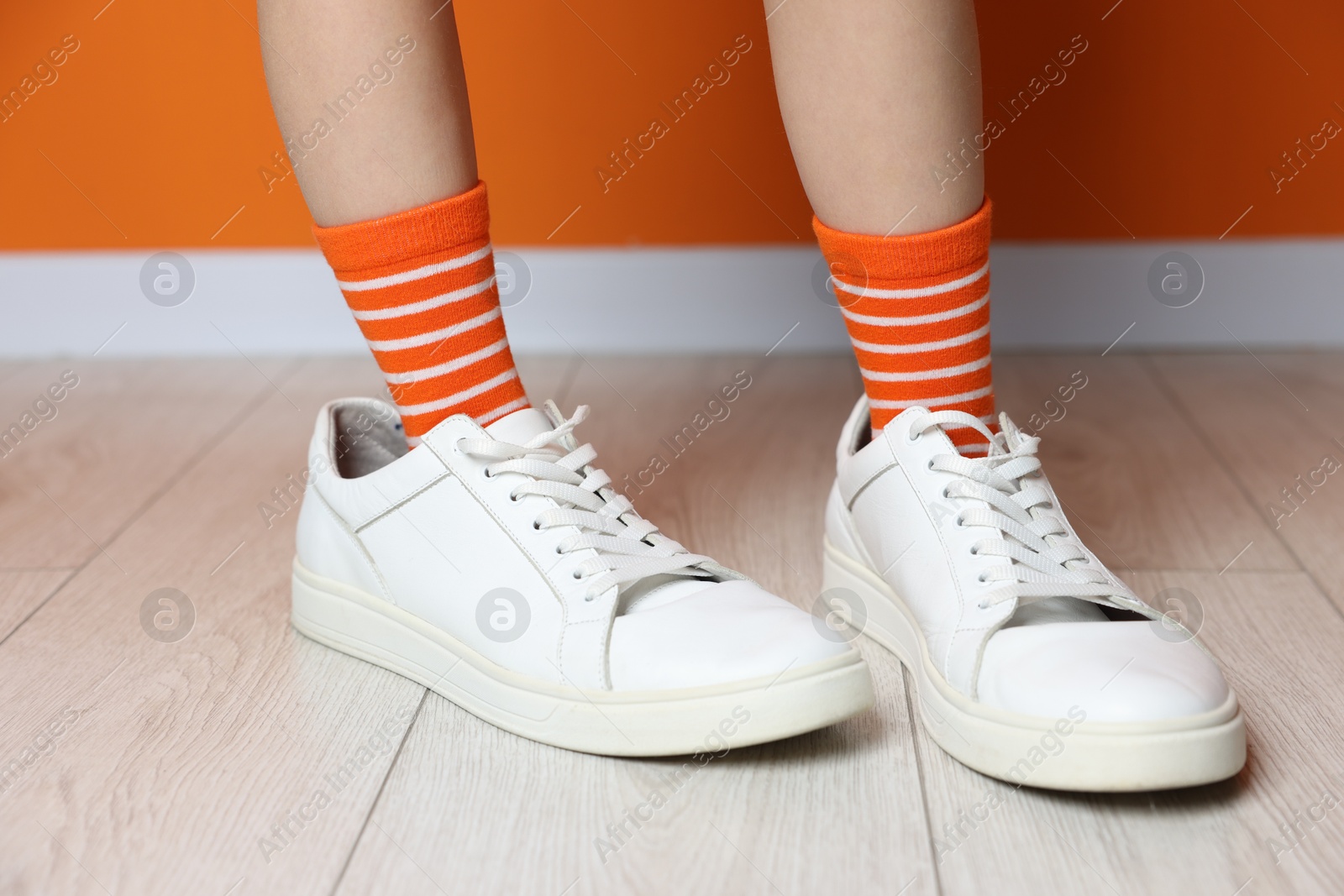 Photo of Child wearing oversized sneakers near orange wall indoors, closeup