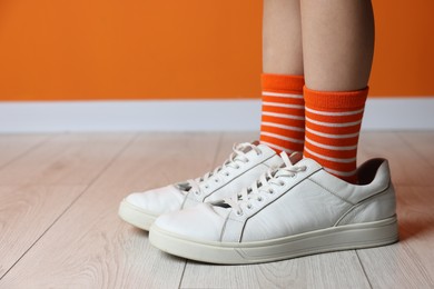 Photo of Child wearing oversized sneakers near orange wall indoors, closeup. Space for text