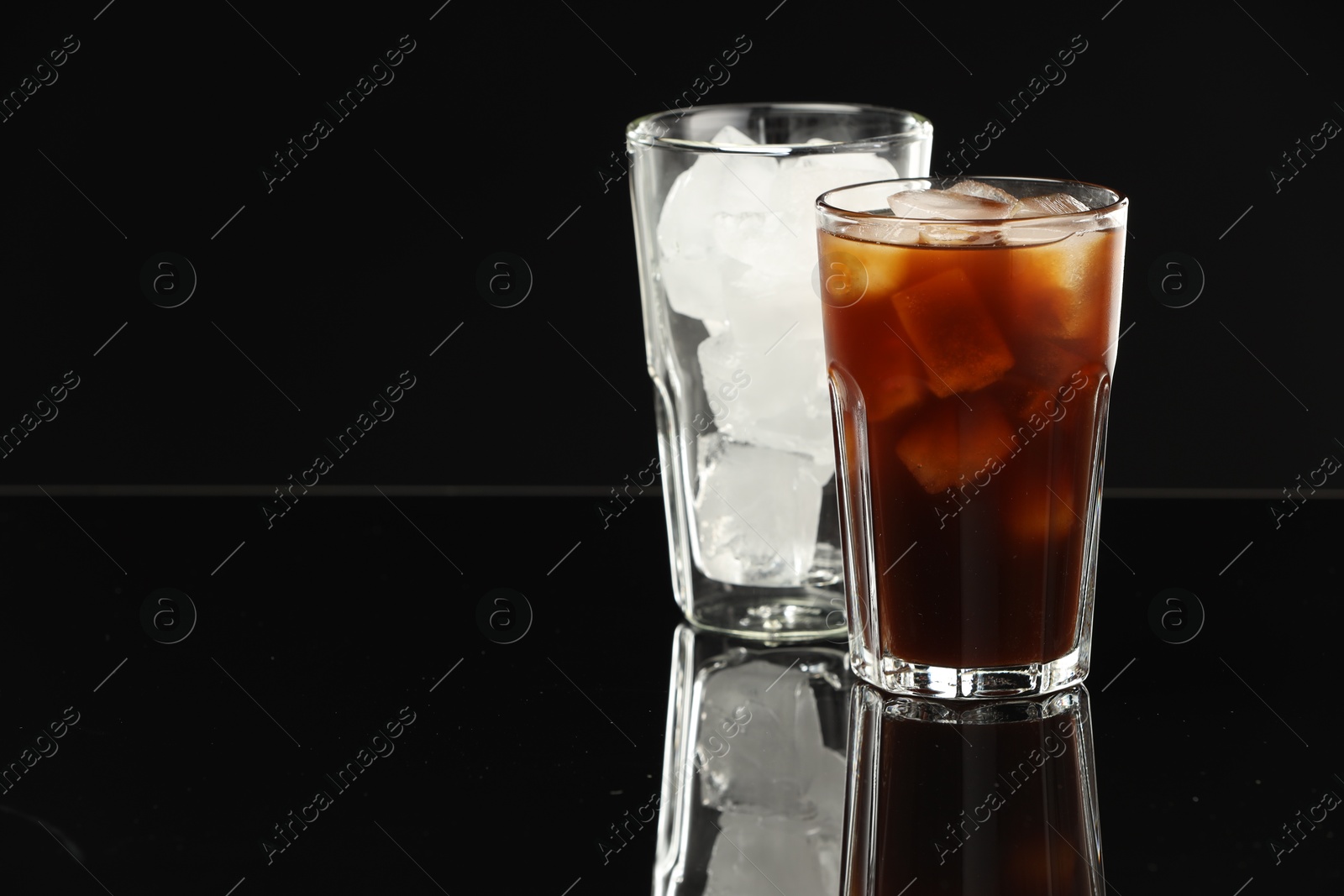 Photo of Refreshing iced coffee and ice cubes in glasses on black mirror surface. Space for text