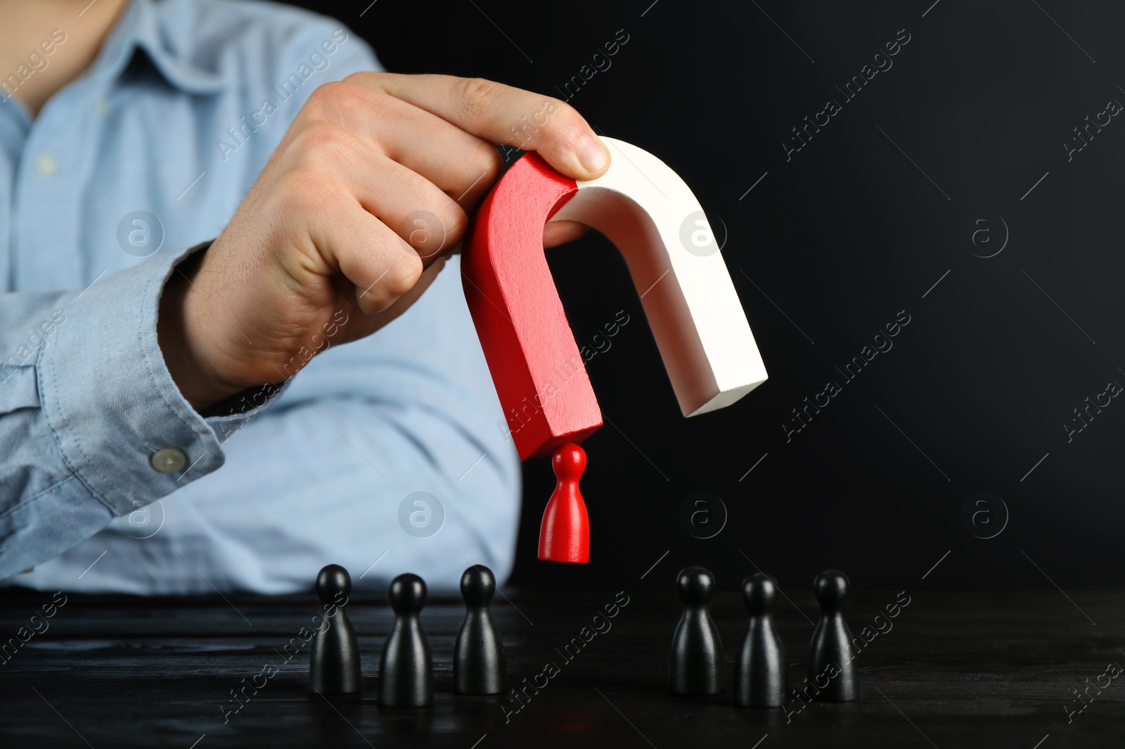 Photo of Man with magnet attracting red piece among black ones at wooden table, closeup