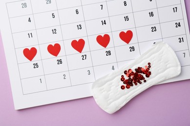 Photo of Calendar with marked dates and menstrual pad with sequins on lilac background, top view