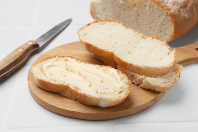 Photo of Fresh bread with butter and knife on white tiled table, closeup