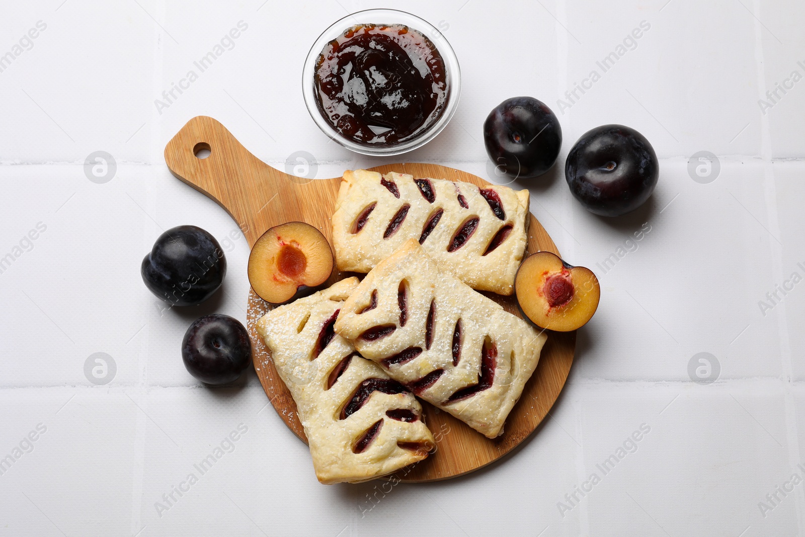 Photo of Delicious puff pastries, plums and jam on white tiled table, top view