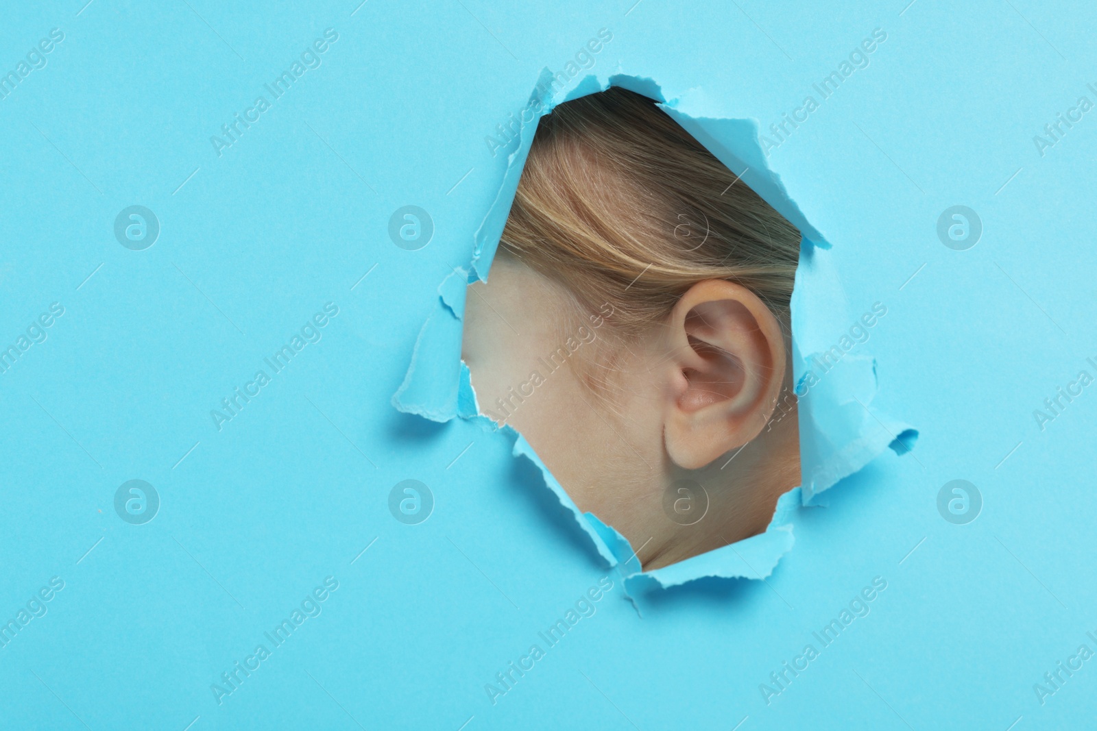 Photo of Little girl showing her ear through hole in light blue paper, closeup. Space for text