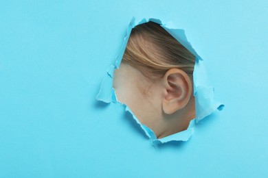 Little girl showing her ear through hole in light blue paper, closeup. Space for text