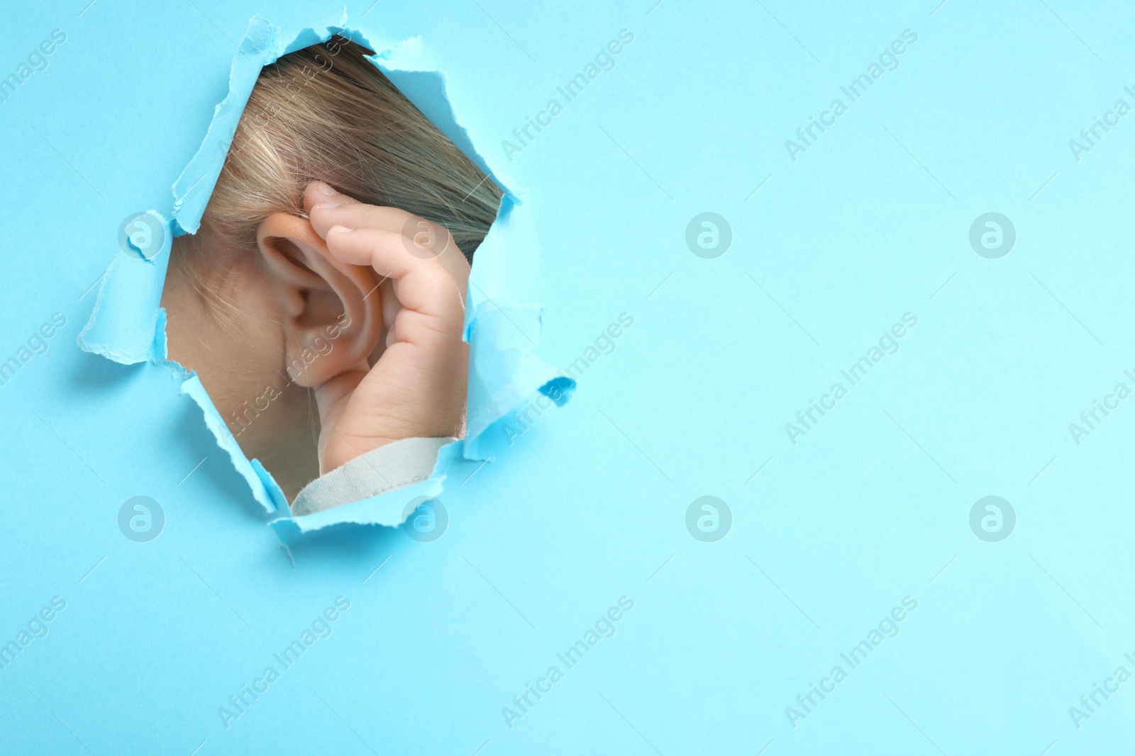 Photo of Little girl showing hand to ear gesture through hole in light blue paper, closeup. Space for text