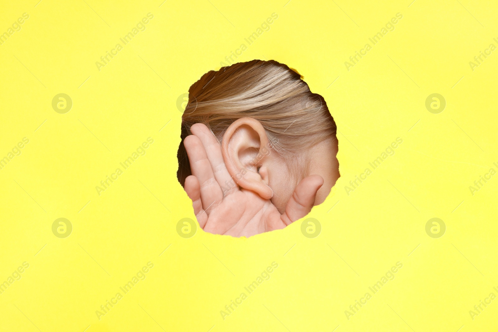 Photo of Little girl showing hand to ear gesture through hole in yellow paper, closeup