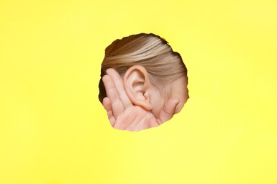 Photo of Little girl showing hand to ear gesture through hole in yellow paper, closeup