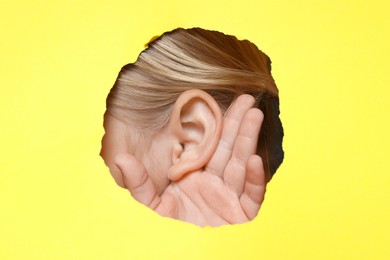 Photo of Little girl showing hand to ear gesture through hole in yellow paper, closeup