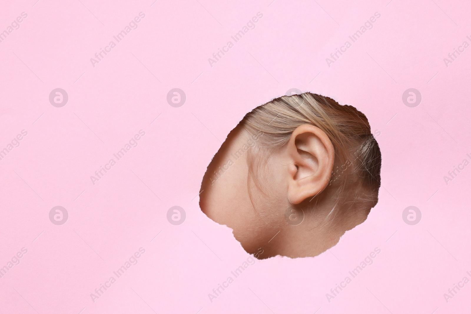 Photo of Little girl showing her ear through hole in pink paper, closeup. Space for text