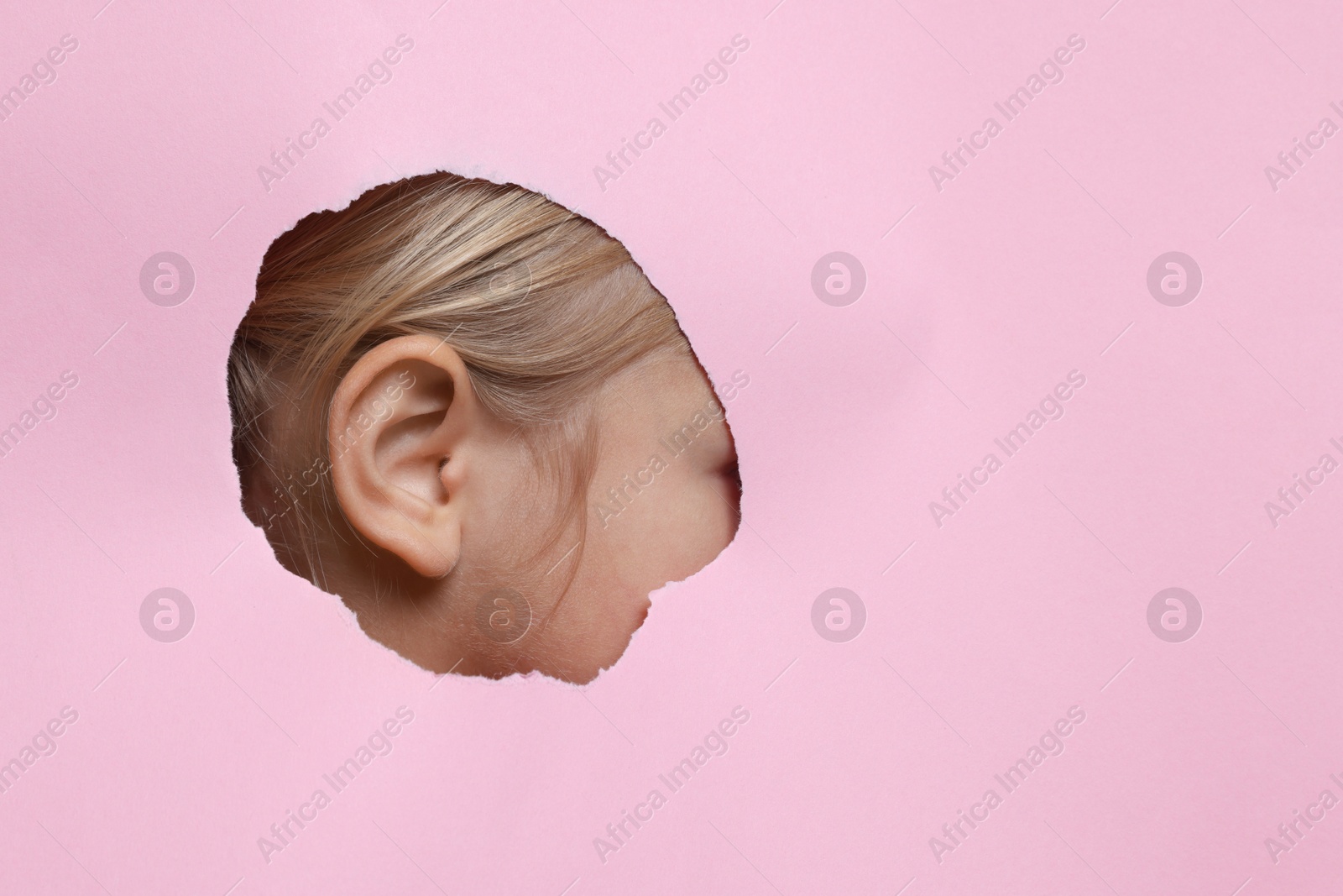 Photo of Little girl showing her ear through hole in pink paper, closeup. Space for text