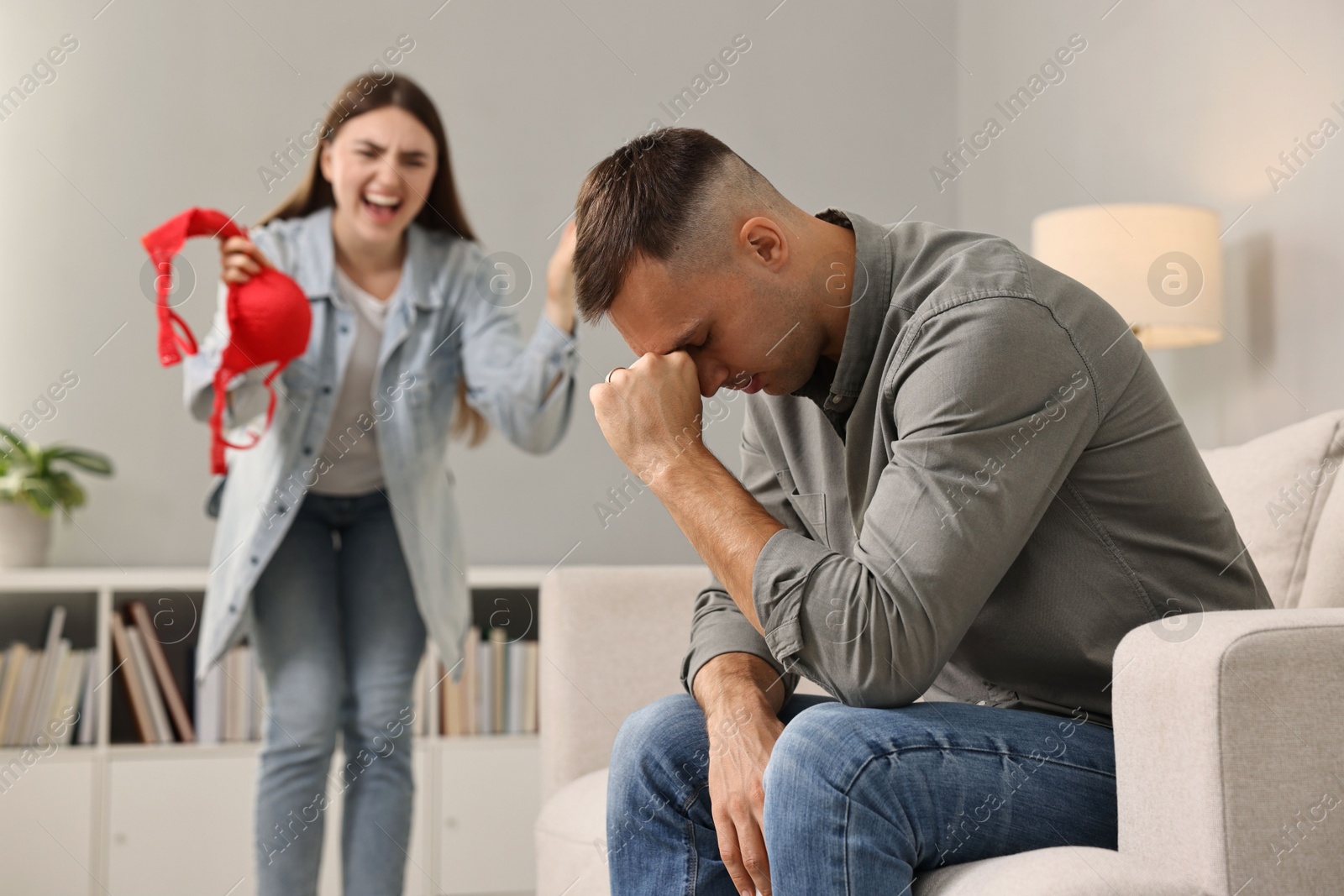 Photo of Emotional wife with mistress bra as proof screaming at her unfaithful husband while he ignoring her indoors, selective focus