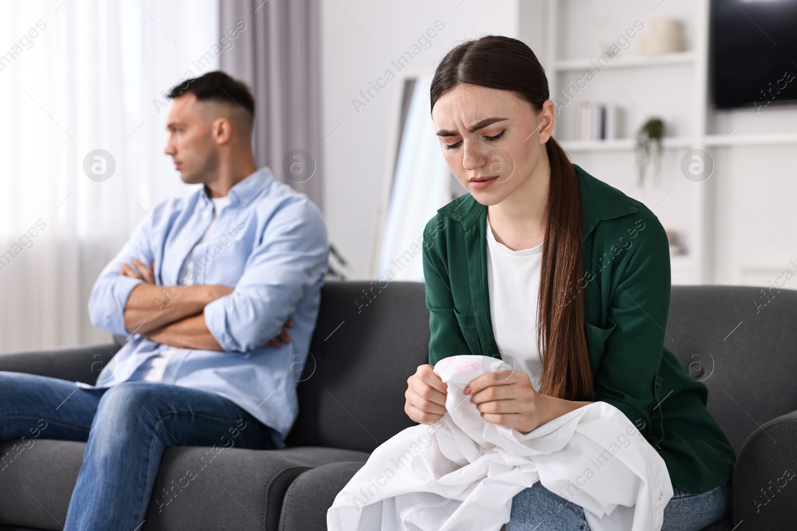 Photo of Frustrated wife holding shirt with lipstick marks as proof of her unfaithful husband at home