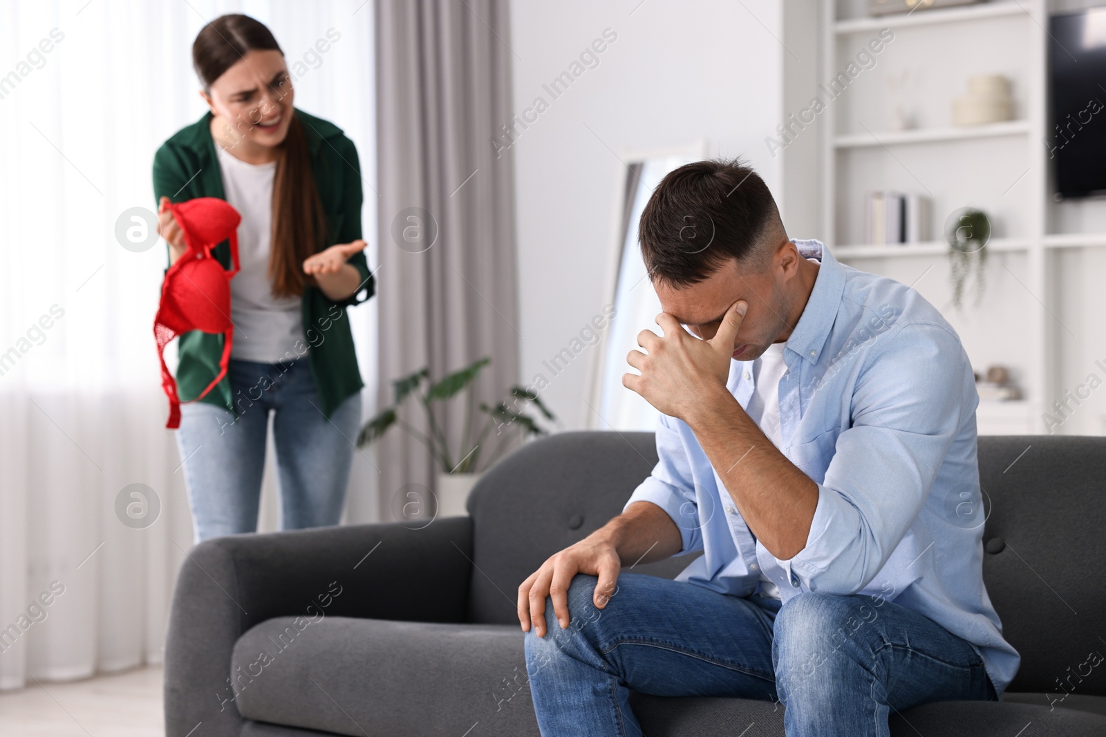 Photo of Emotional wife with mistress bra as proof screaming at her unfaithful husband while he ignoring her indoors, selective focus